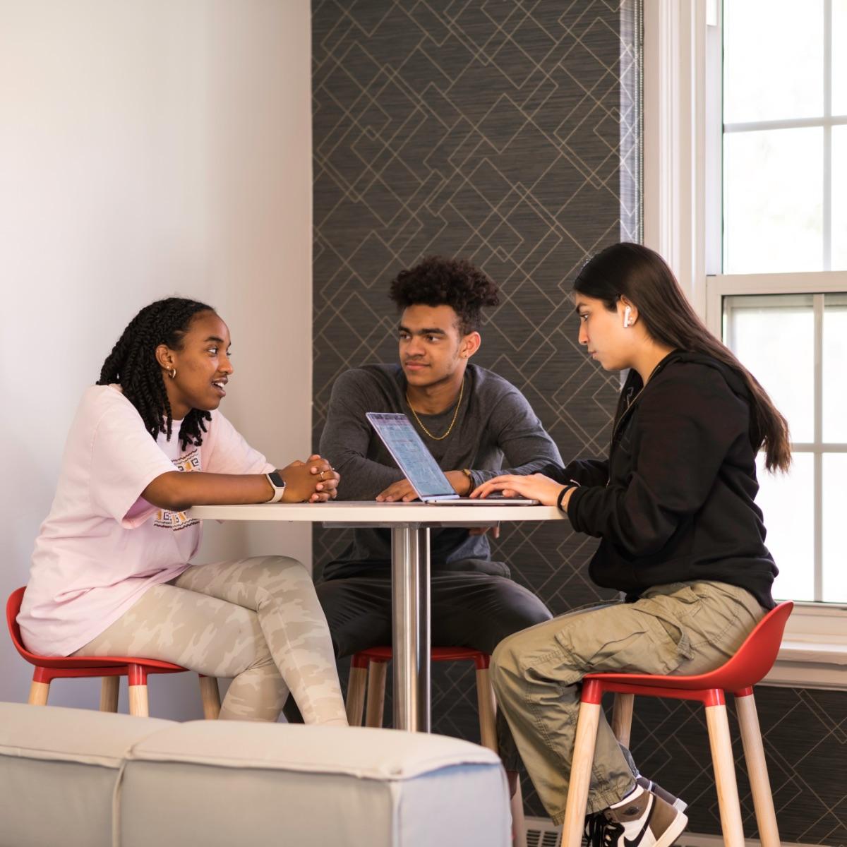 Three students sitting at a round desk. One looks at their laptop